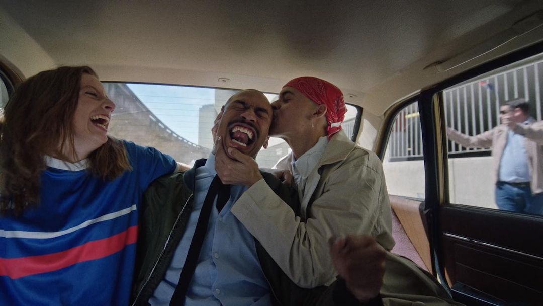 Three adults laugh together inside of a car in the city with buildings visible in the background from the car windows.