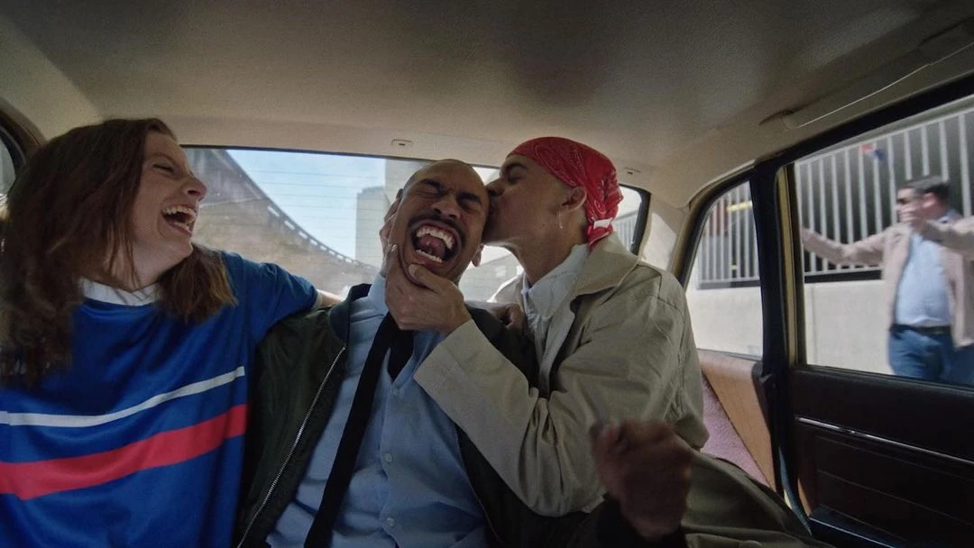Three adults laugh together inside of a car in the city with buildings visible in the background from the car windows.