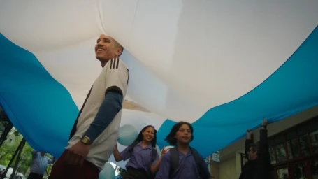 Smiling man in sports jersey and children holding a large white and blue banner outdoors directed by Lana Senaoui