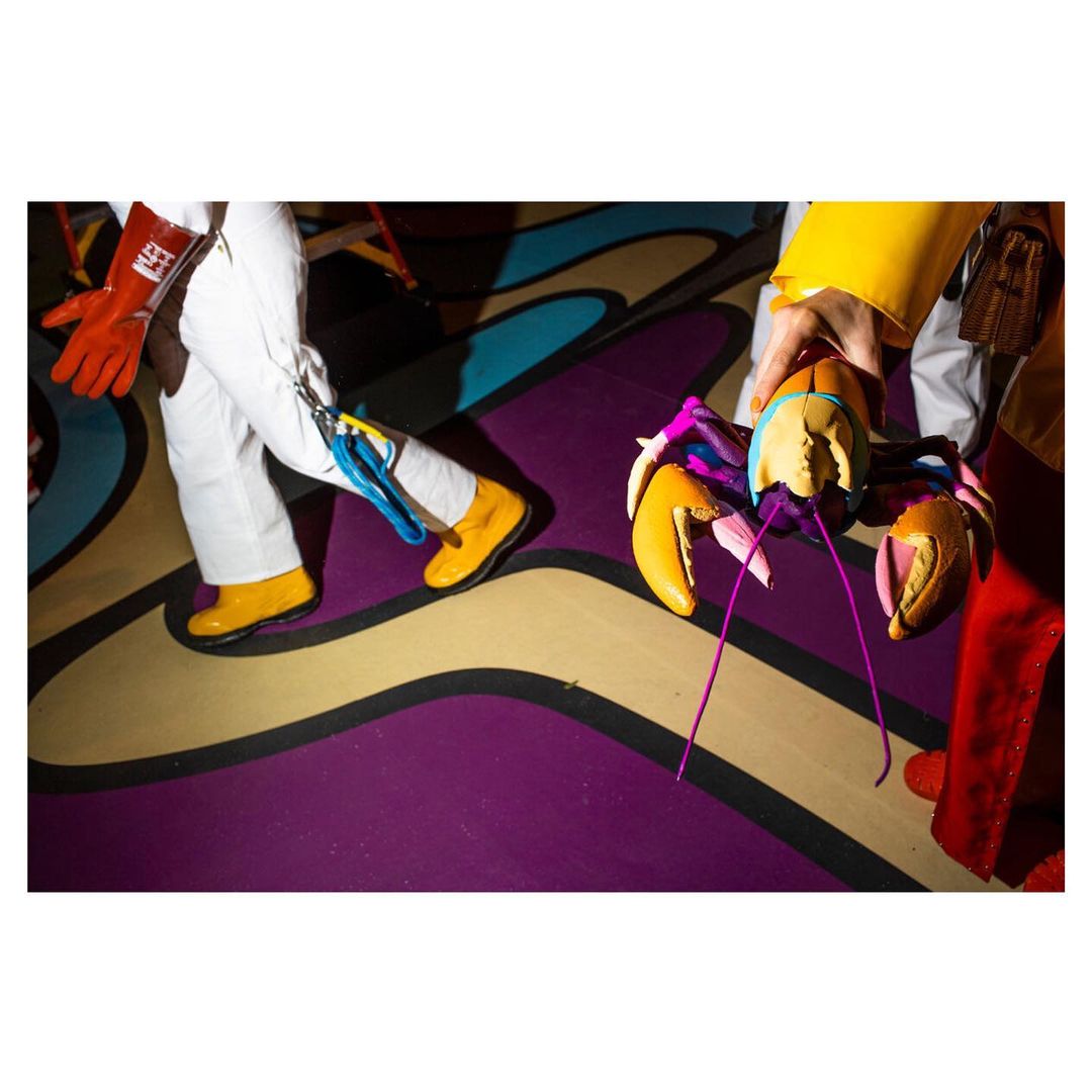 Two people in white and yellow clothing hold a colorful lobster puppet on a floor with purple and yellow swirling designs.