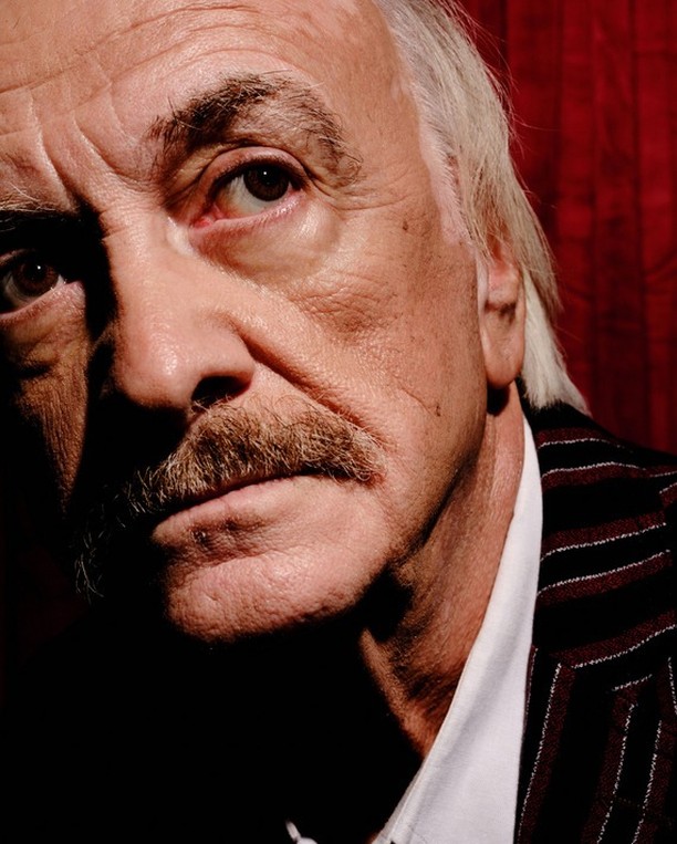 Portrait of an elderly adult male with a mustache and white hair, wearing a striped jacket, against a dark red curtain backdrop.