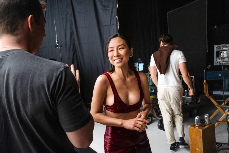 Smiling Asian woman in red outfit with film crew in the background at a photo studio with lighting equipment and black backdrop.