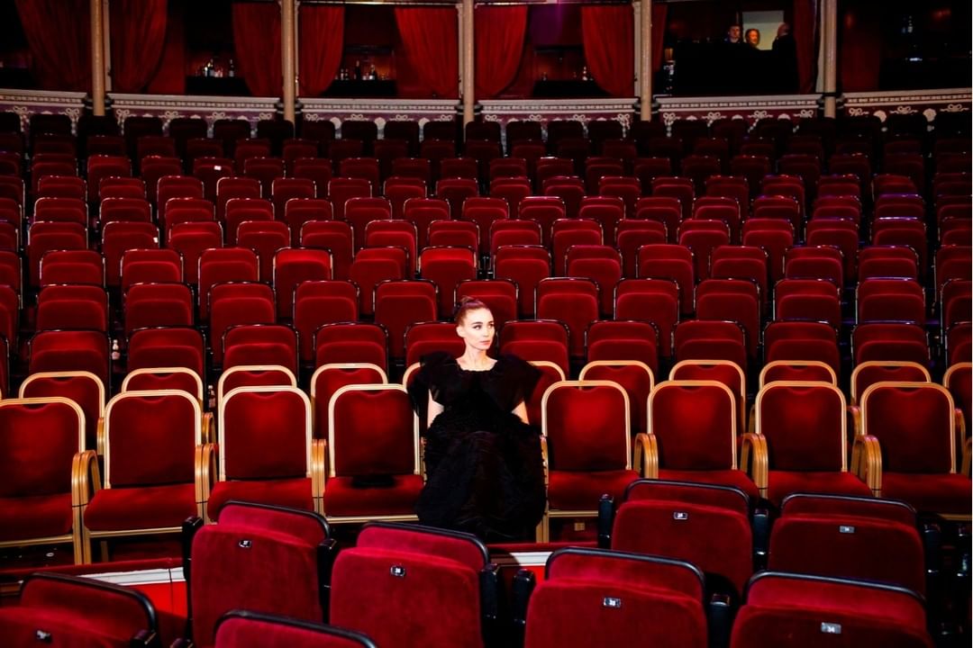 A woman in a black dress sits in an otherwise empty theater. Red velvet seats fill the auditorium, creating a stark contrast.