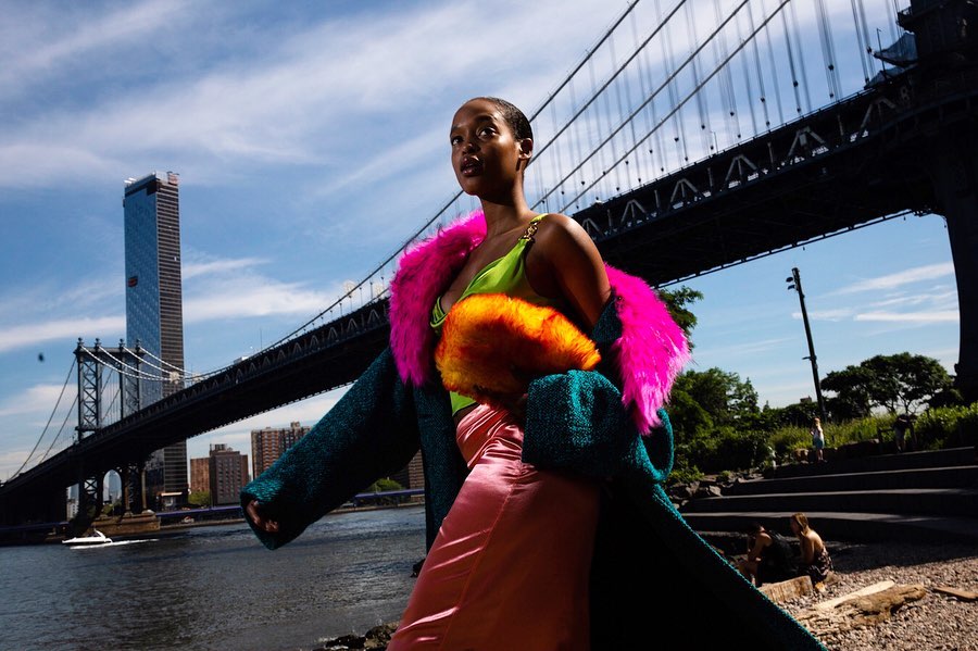A fashionable woman poses near the Manhattan Bridge wearing a teal coat with pink fur trim, a neon green top, and a peach skirt.