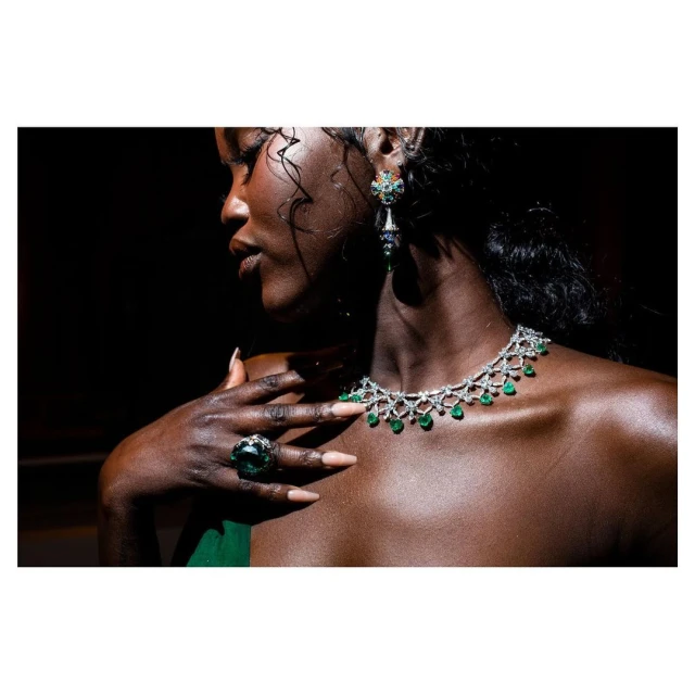 Elegant woman with jewelry: Close-up of a dark-skinned woman wearing emerald necklace, ring, and earrings, with a blurred black backdrop.