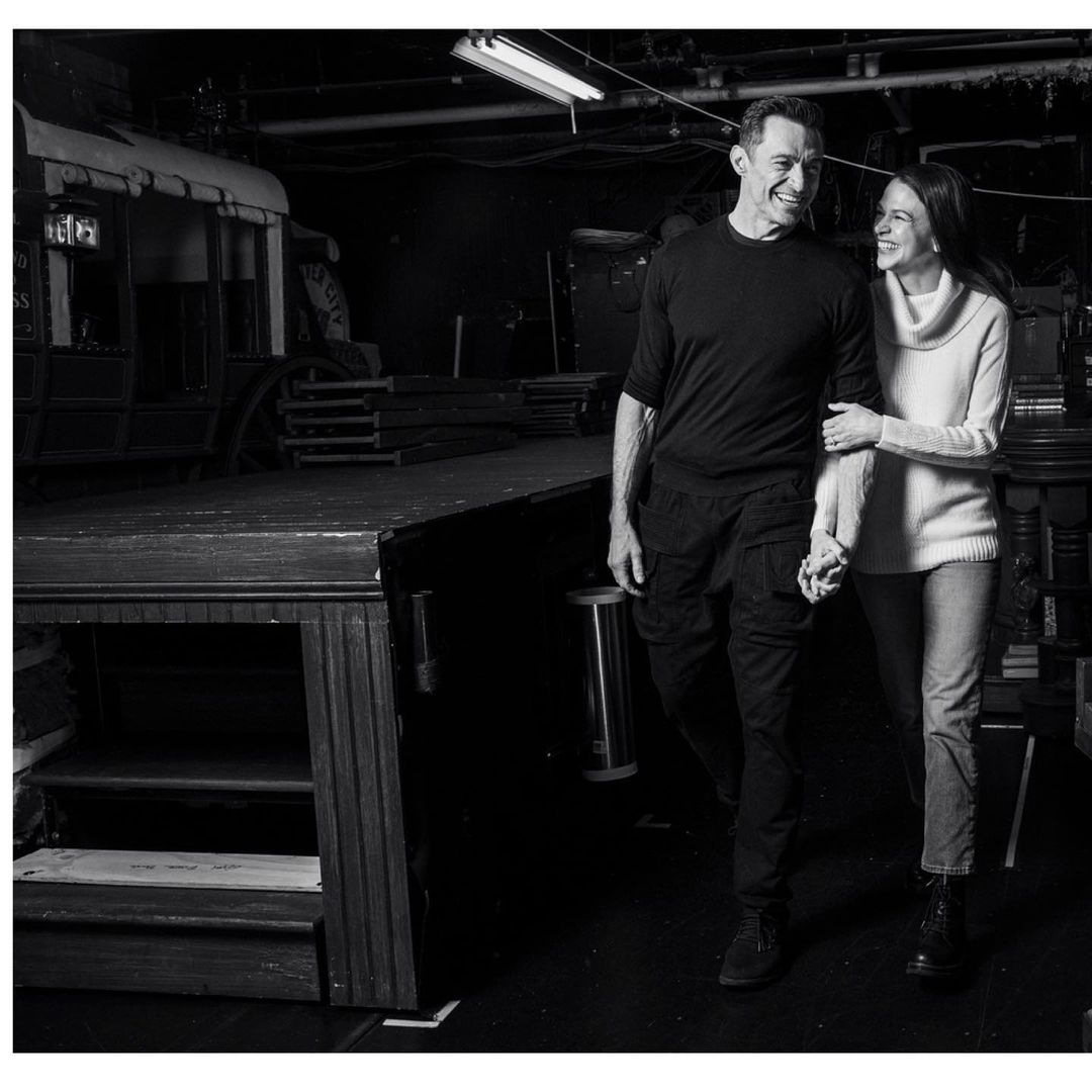 Black and white shot of a smiling couple holding hands, walking through a backstage area with theater props and stage equipment.