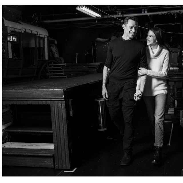 Black and white shot of a smiling couple holding hands, walking through a backstage area with theater props and stage equipment.