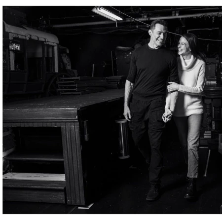 Black and white shot of a smiling couple holding hands, walking through a backstage area with theater props and stage equipment.