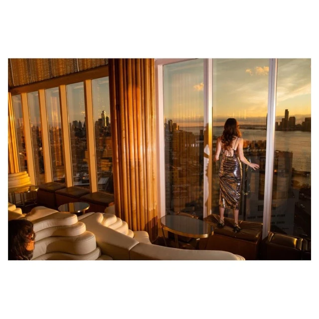 Woman in shiny dress stands at the window of a high-rise, overlooking a cityscape at sunset. Interior view with modern lounge seating.