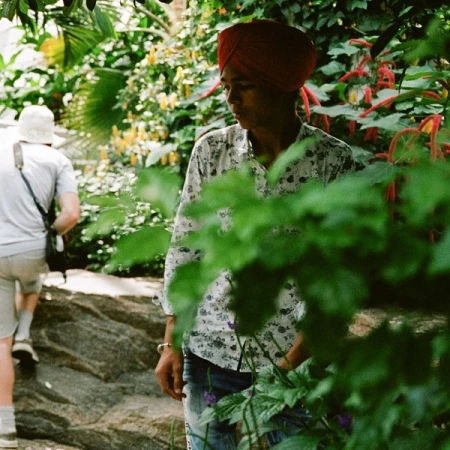 Man in turban and floral shirt stands in a lush garden with another person visible in the background walking along a stone path.
