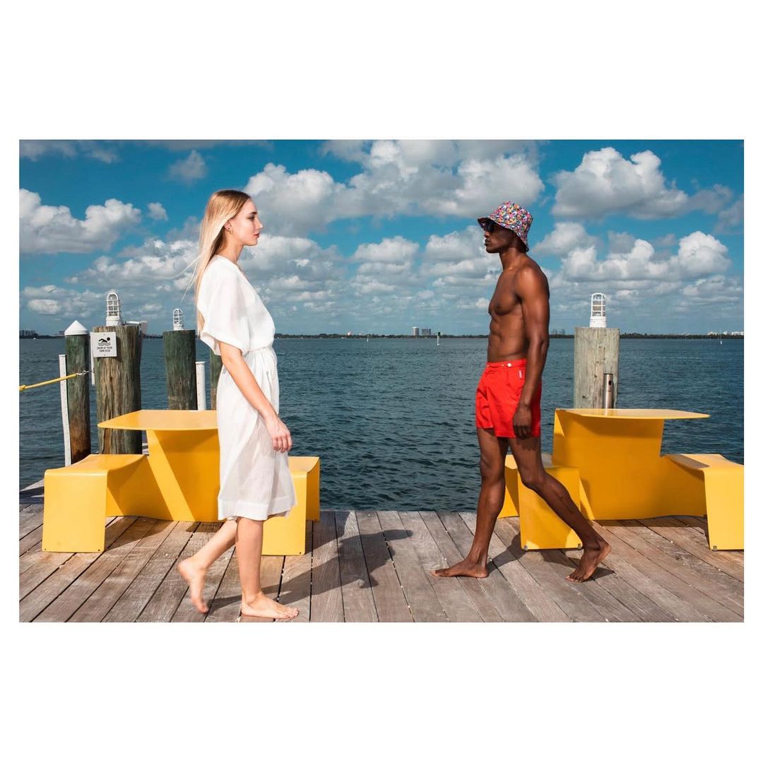 Two people walk on a wooden pier near yellow benches with the ocean and blue sky in the background, creating a stylish, outdoor scene.