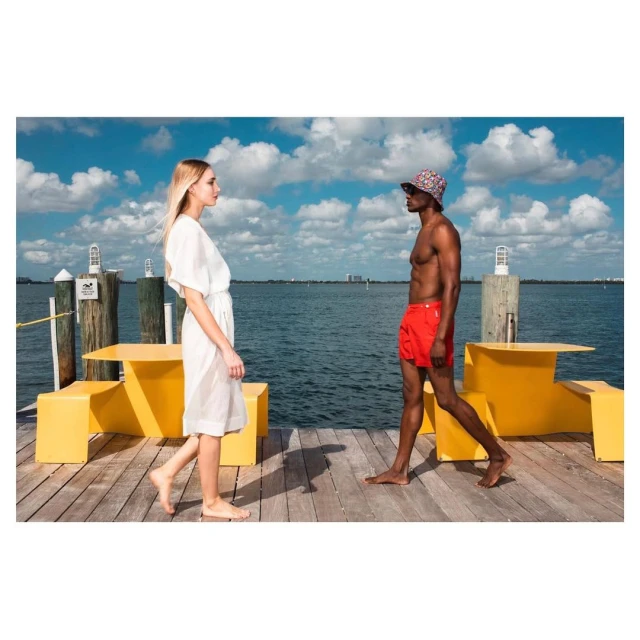 Two people walk on a wooden pier near yellow benches with the ocean and blue sky in the background, creating a stylish, outdoor scene.