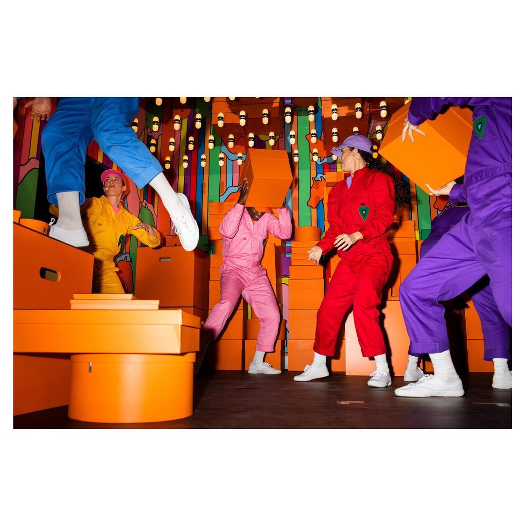 Group of five people in colorful jumpsuits holding orange boxes in a brightly lit, surreal-looking indoor environment.