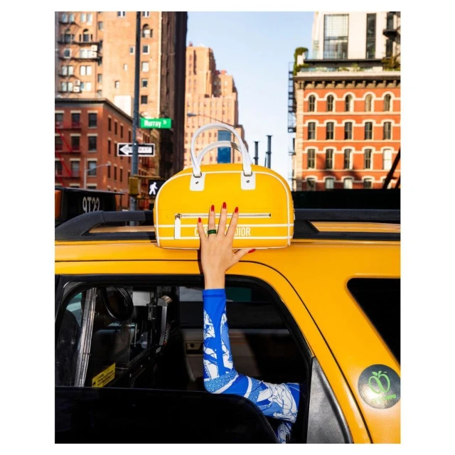Bright yellow handbag displayed from taxi in New York City, featuring red manicured hand, with cityscape buildings in the background.