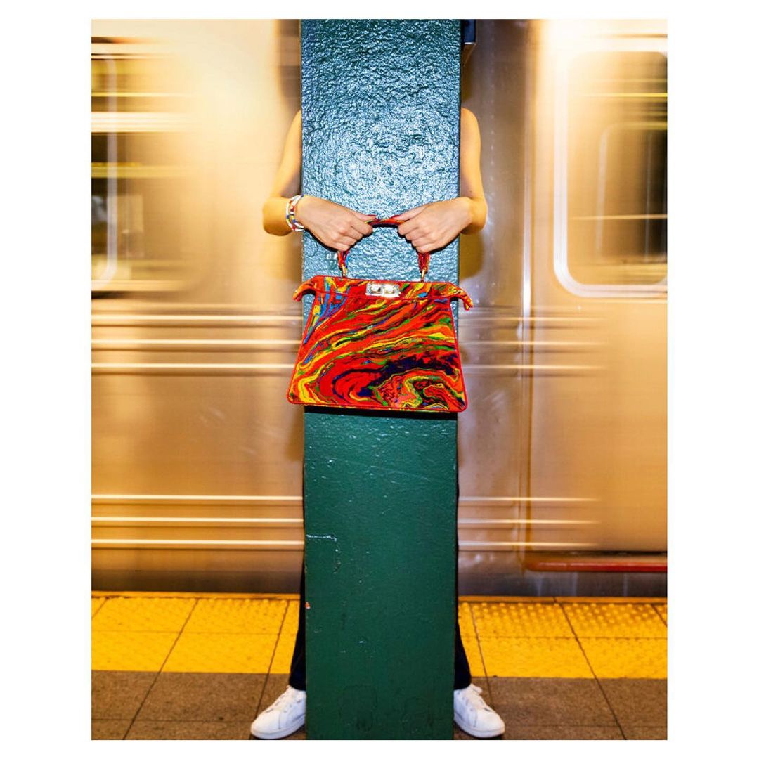 Colorful handbag held by person partially obscured by green column against blurred train in subway station, white sneakers visible.