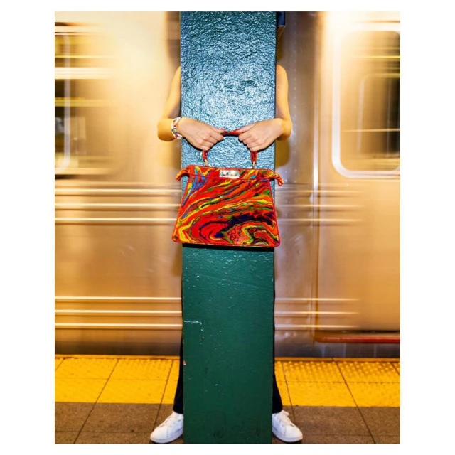 Colorful handbag held by person partially obscured by green column against blurred train in subway station, white sneakers visible.