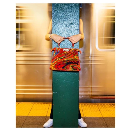 Colorful handbag held by person partially obscured by green column against blurred train in subway station, white sneakers visible.