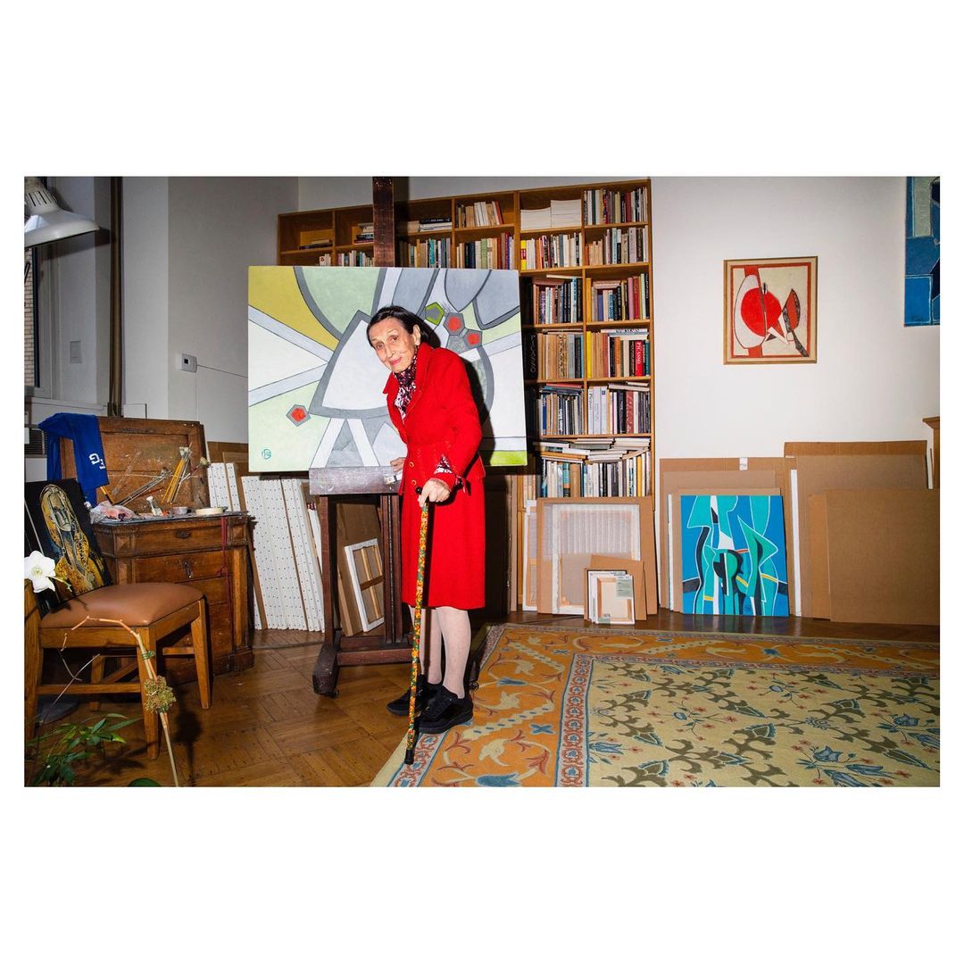 Elderly woman in red coat with cane stands in an artist studio, painting on an easel, surrounded by paintings and bookshelves.