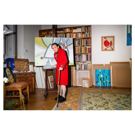 Elderly woman in red coat with cane stands in an artist studio, painting on an easel, surrounded by paintings and bookshelves.