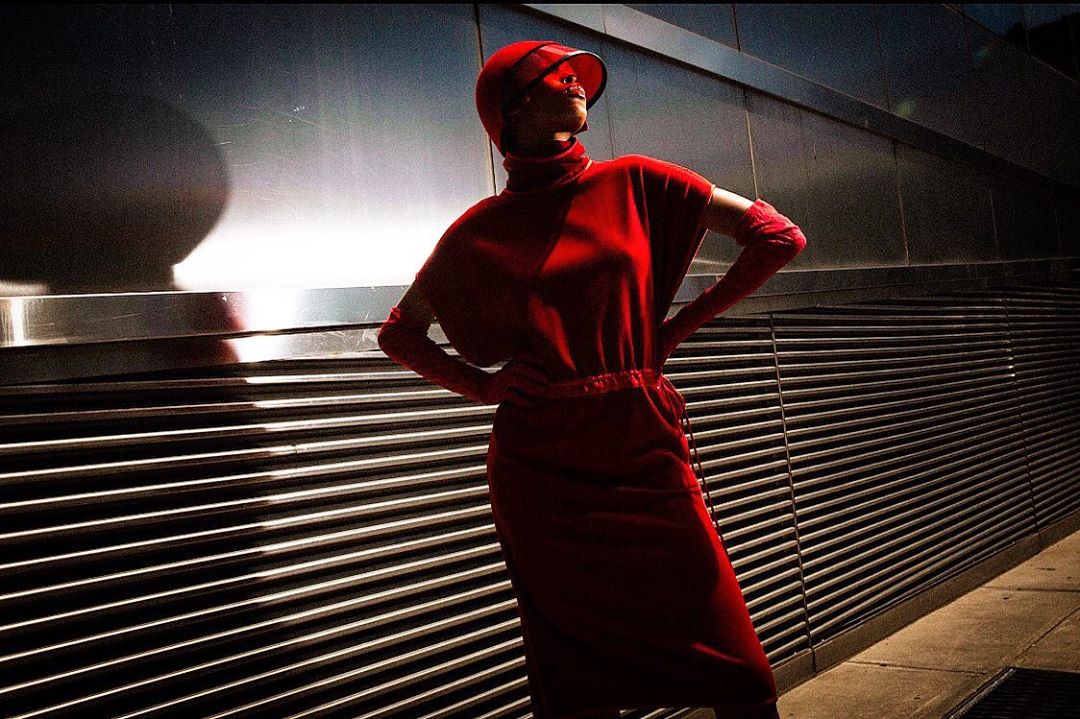 Fashionable adult female in a red dress and headwear posing in front of a textured wall with shadow and light contrast. Urban environment.
