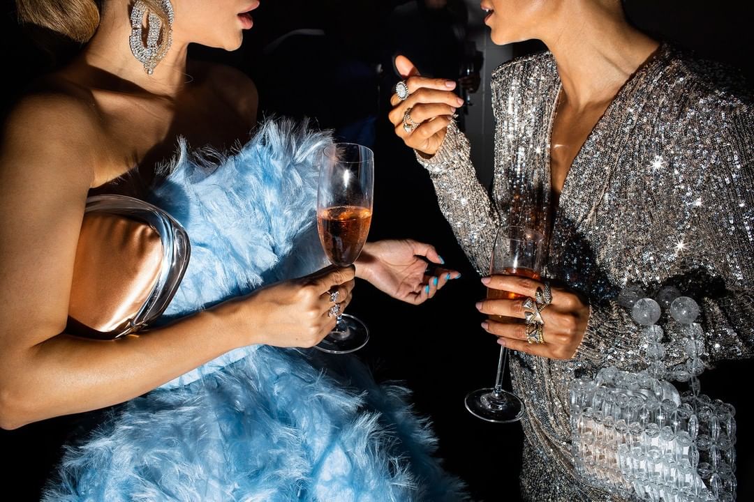 Two women in glittering gowns toast with champagne at a party. The woman on the left wears a light blue dress with feather details.