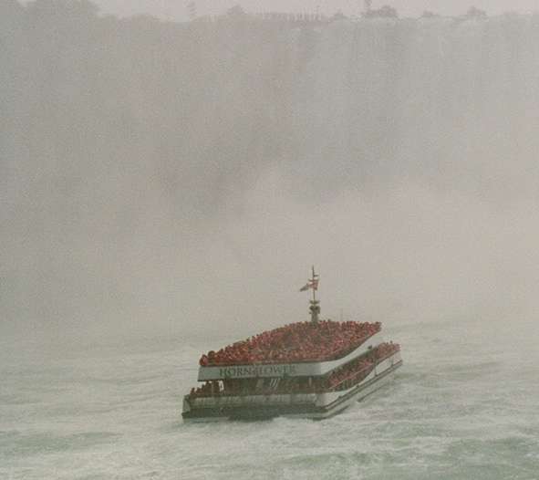 Large tour boat crowded with people navigates choppy water near Niagara Falls amidst a hazy mist, emphasizing travel and tourism.