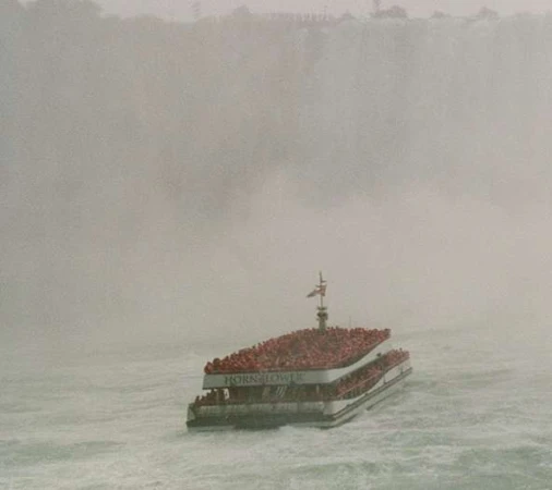 Large tour boat crowded with people navigates choppy water near Niagara Falls amidst a hazy mist, emphasizing travel and tourism.