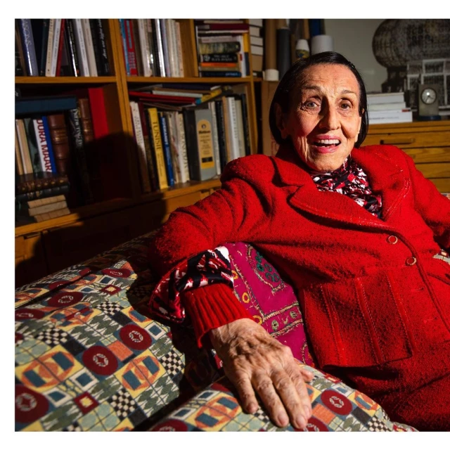 Elderly woman in a red suit smiles while sitting on a patterned couch in front of a bookshelf filled with books indoors.
