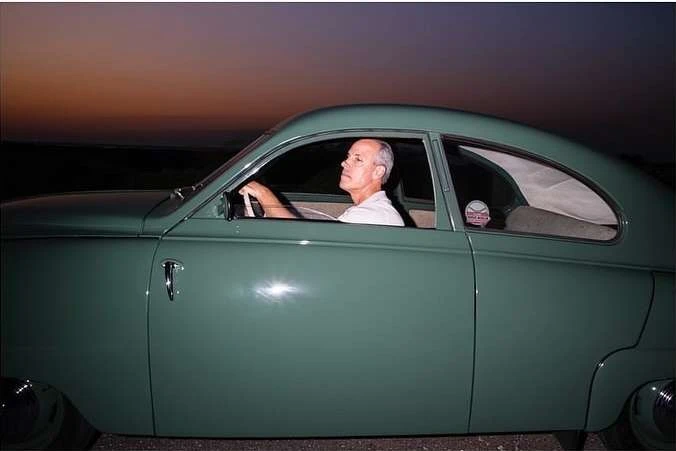 Adult male driving a vintage green Saab car, profile view, evening light, rural setting, classic car enthusiast lifestyle, automotive.