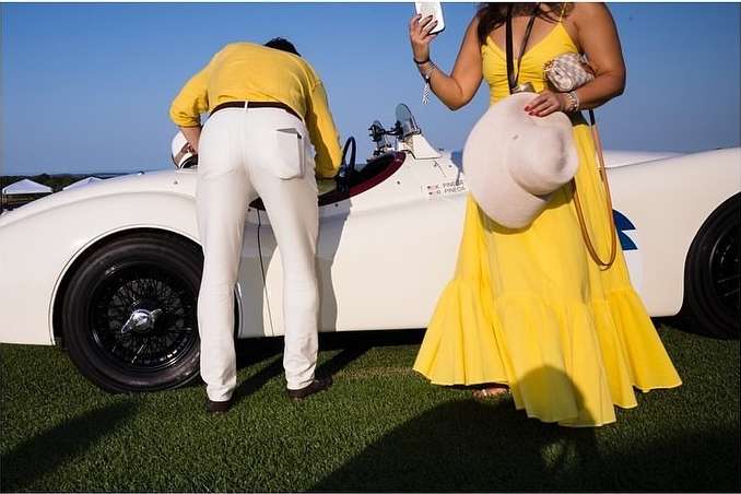 Woman in yellow dress holds hat near a classic white car with a man in white pants and yellow shirt on a sunny day.