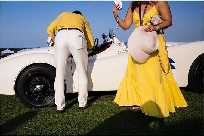 Woman in yellow dress holds hat near a classic white car with a man in white pants and yellow shirt on a sunny day.