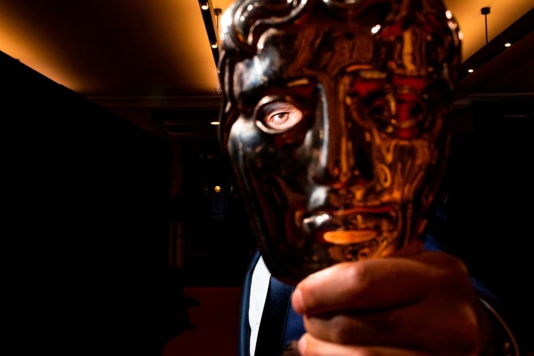 Close-up studio shot of a person holding a gold-colored mask in front of their face, partially obscuring their features in dark lighting.