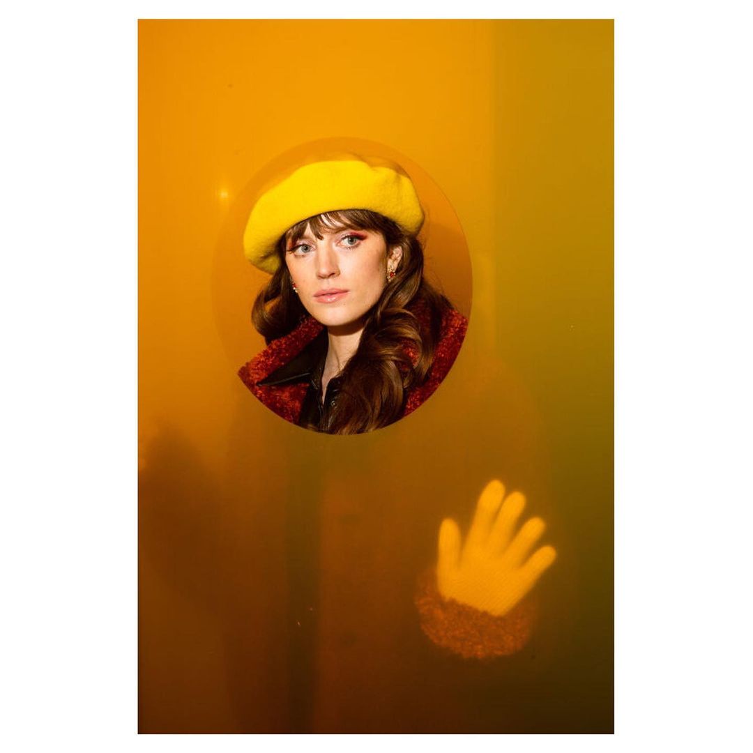 Portrait of a woman with brunette hair and a yellow beret looking through a round opening in a yellow surface with a hand below.