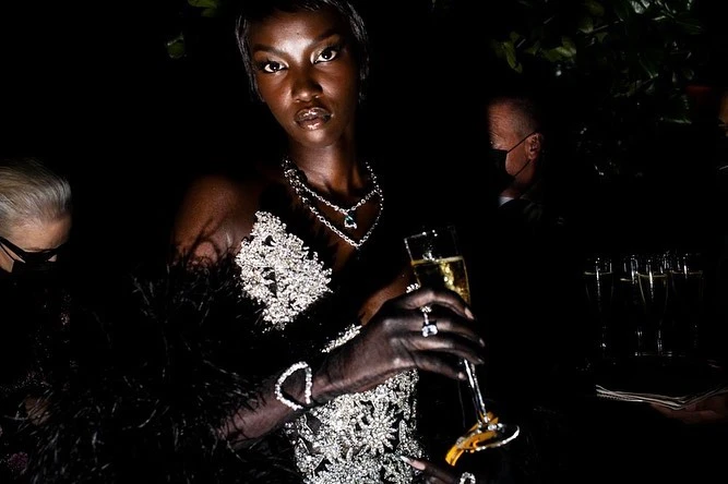 Elegant African woman in embellished gown holds champagne at evening event with muted background guests and subtle natural lighting.