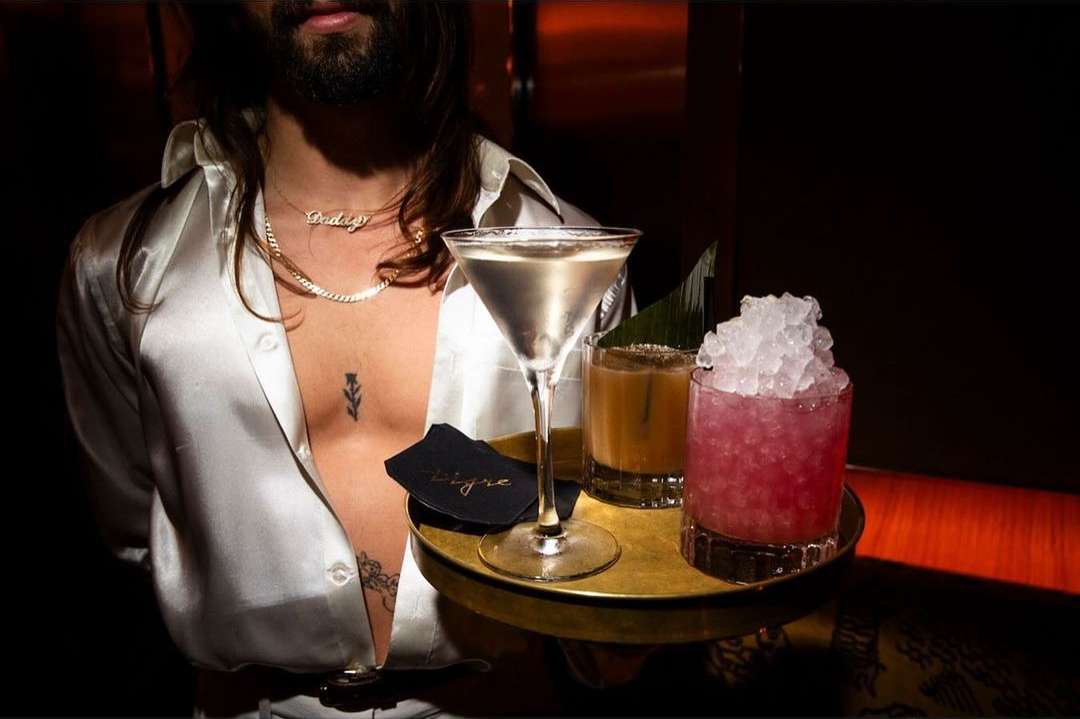 Bartender in white shirt serving cocktails on gold tray with martini, pink cocktail, and amber drink in dark lounge setting.