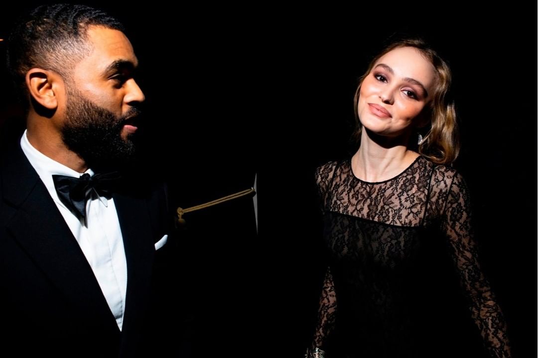 A man in a tuxedo and a woman in a lace dress pose against a dark backdrop, illuminated by spotlights in an elegant portrait.