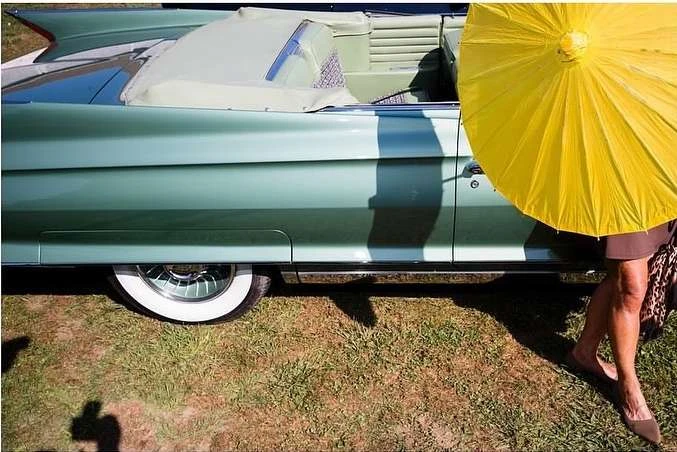 Vintage turquoise convertible car parked on grass, partially shaded by a yellow umbrella and standing person on the right.