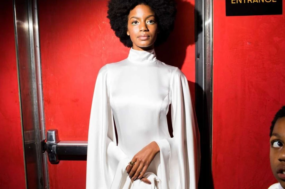 Fashion portrait of a Black woman in white dress standing by a red door, with a child partially visible on the right side.