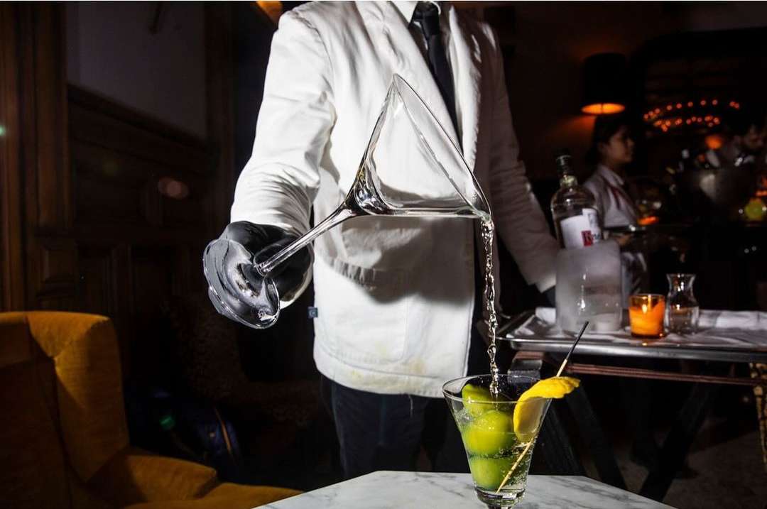 Bartender pours clear liquid from a martini glass into a cocktail glass with a lemon garnish, against a dimly lit bar backdrop.