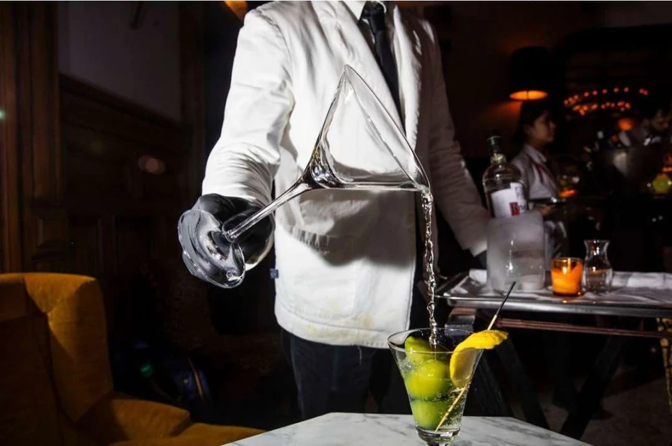 Bartender pours clear liquid from a martini glass into a cocktail glass with a lemon garnish, against a dimly lit bar backdrop.