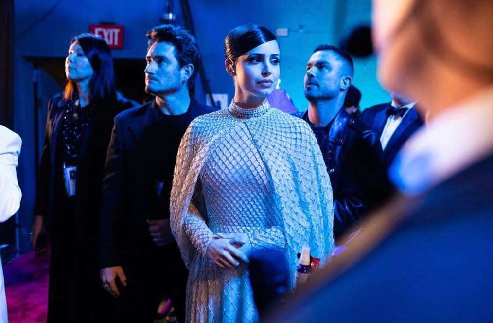 A woman in an elaborate dress stands at an event with three people behind her, all illuminated in blue lighting. 