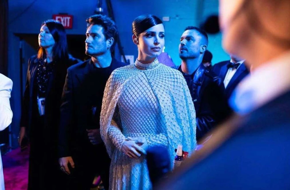 A woman in an elaborate dress stands at an event with three people behind her, all illuminated in blue lighting. 