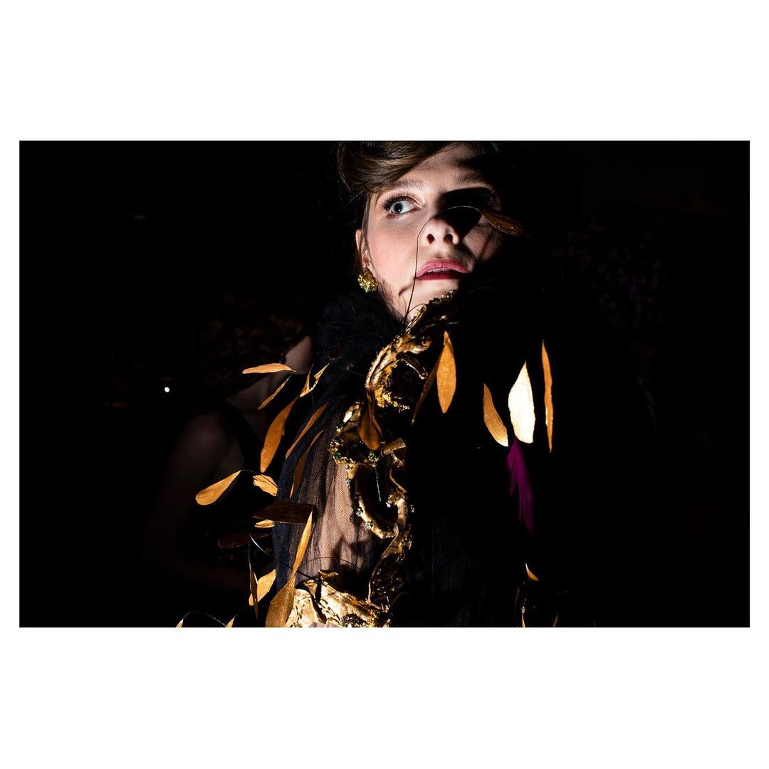 Woman wearing a feather boa with gold leaf embellishments, posing against a dark backdrop in a close-up fashion shot.