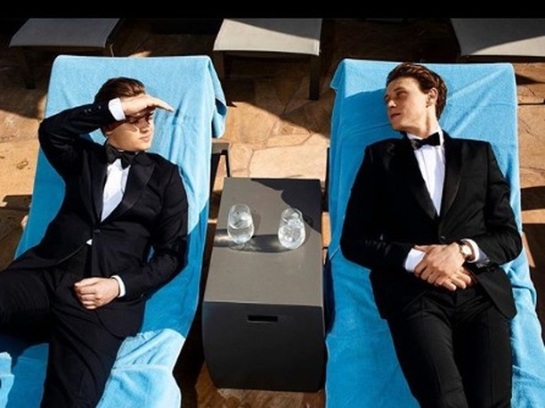 Two men in formal wear lounge on poolside chairs with drinks, basking in the sunlight reflecting off the stone patio.