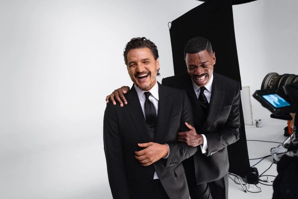 Two smiling adult males in black suits pose in a photo studio, captured at eye-level with visible camera equipment, and a plain white backdrop.