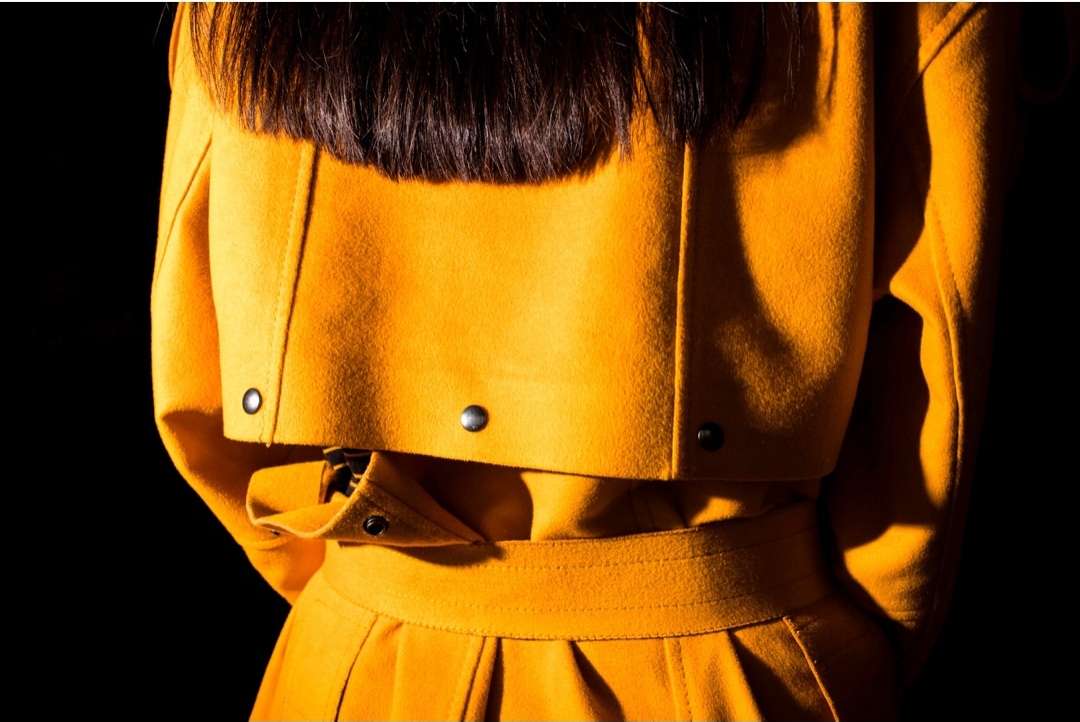 A woman with dark hair wears a cropped mustard-yellow wool jacket and skirt set against a stark black background in this fashion-forward image.