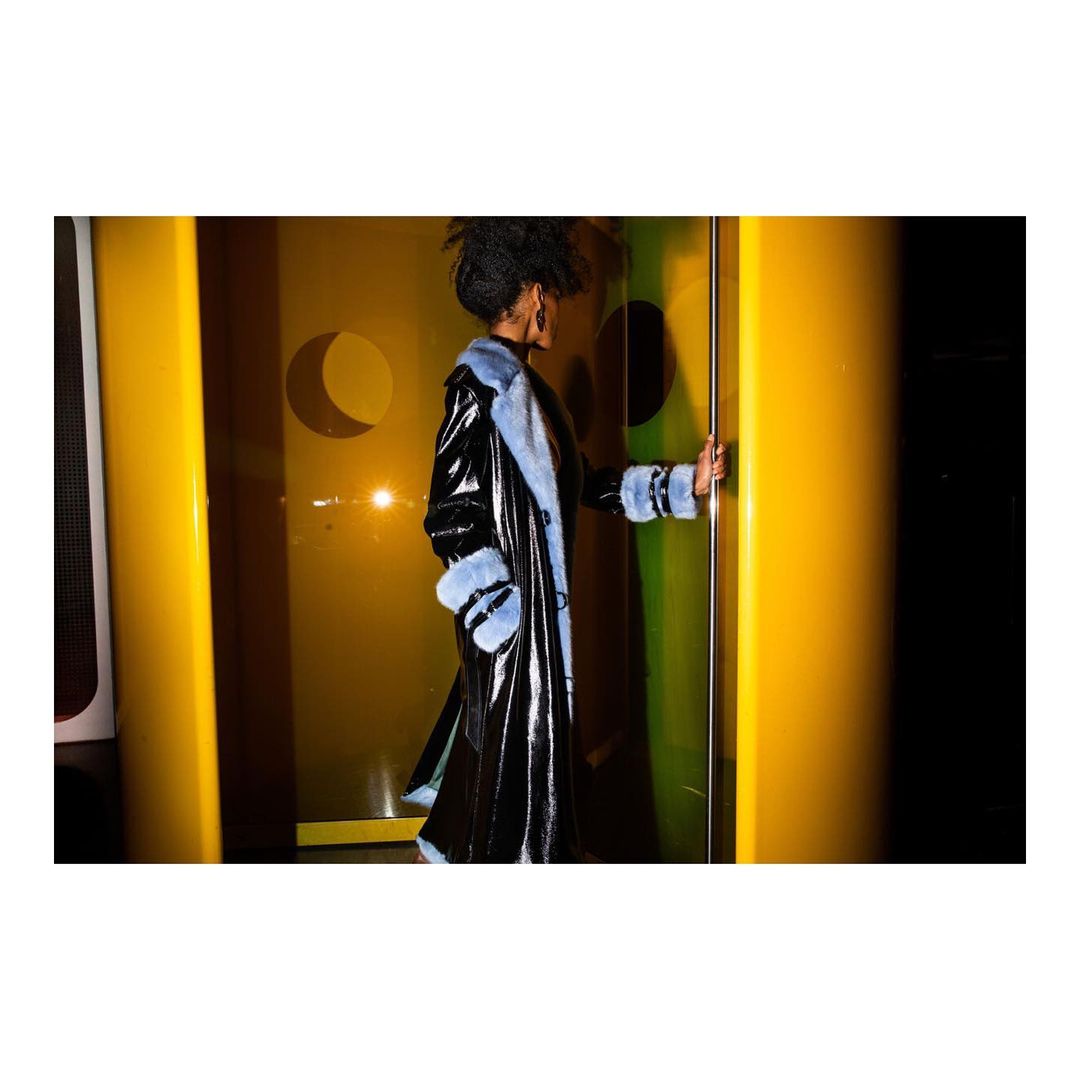 Woman in a shiny black coat with light blue fur trim standing in a yellow-lit doorway. Fashion shot, studio lighting.