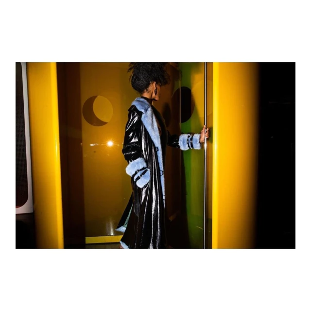 Woman in a shiny black coat with light blue fur trim standing in a yellow-lit doorway. Fashion shot, studio lighting.