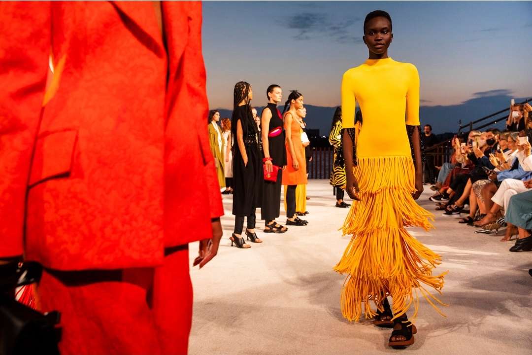 Fashion show on outdoor runway with a Black female model in a yellow dress and a crowd of spectators in the background.