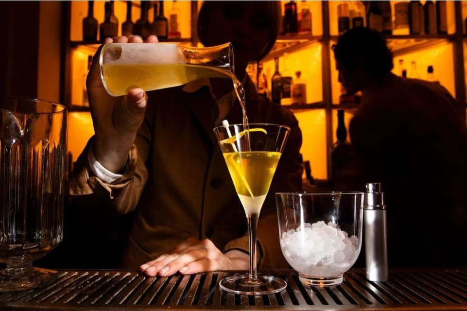 Bartender prepares a cocktail at a bar, pouring from a glass pitcher into a stemmed martini glass; warm lighting, elegant setting.
